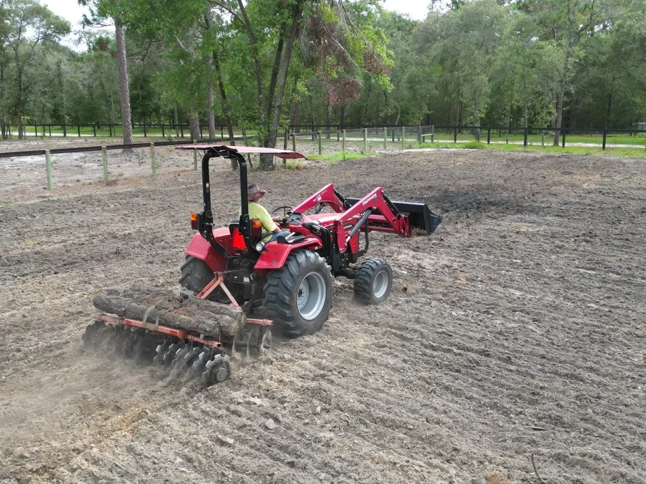 Red compact tractor with front-end loader and disc harrow grading arena soil