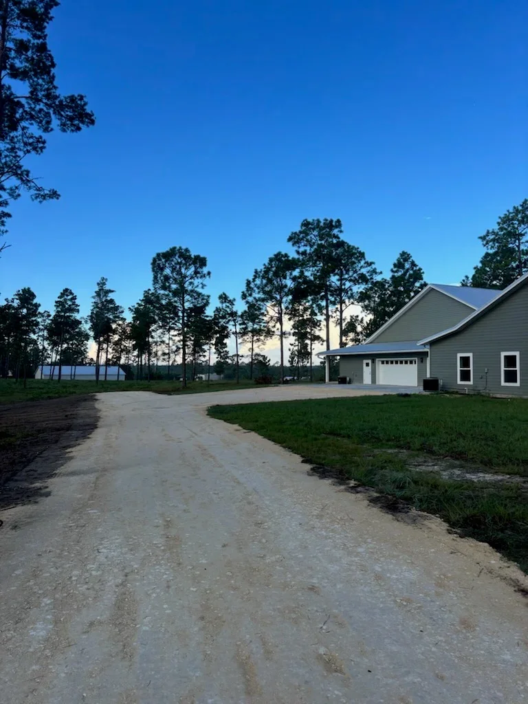 Cleared residential lot with gravel driveway, new home with garage, and pine trees
